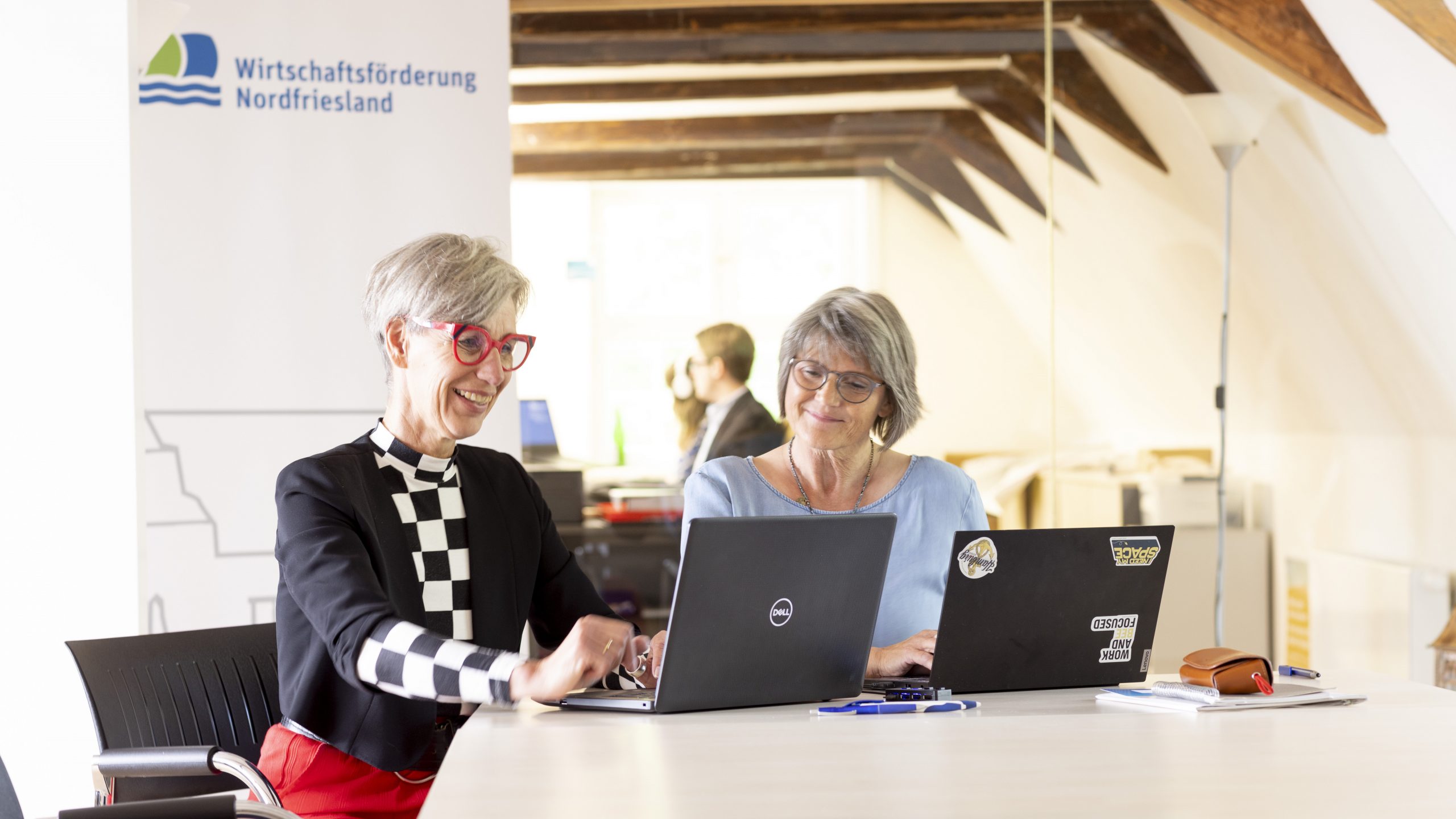 Zwei Teamkolleginnen der Wirtschaftsförderung Nordfriesland sitzen fröhlich nebeneinander vor ihren Laptops.